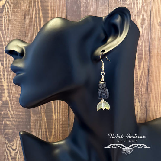 A black cat on a crescent moon earring displayed on a black mannequin ear with 'Nichole Anderson Designs' branding.