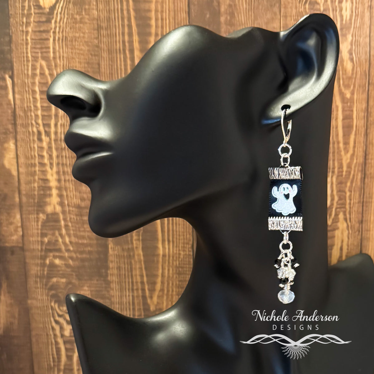 Ghost Ribbon Earring with crystal drops suspended below as drops on a black mannequin ear with 'Nichole Anderson Designs' branding against a wooden background.
