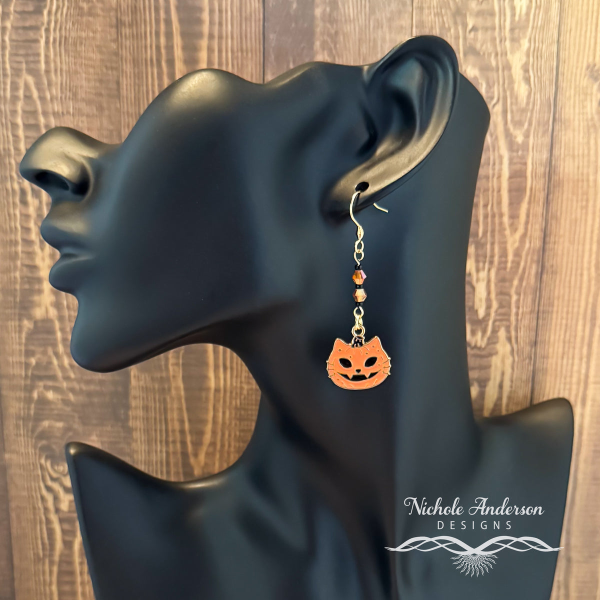 Halloween-themed cat pumpkin jack o lantern earrings on a black mannequin ear with 'Nichole Anderson Designs' branding.