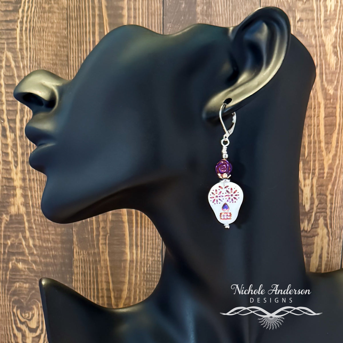 Skull Earring with metallic rose on a black mannequin ear with a wooden background, featuring the brand 'Nichole Anderson Designs'.