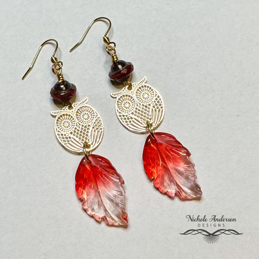Gold owl-shaped earrings with red leaf-like pendants on a light gray background, featuring 'Nichole Anderson Designs' branding.