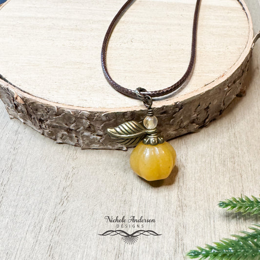 A close up view of a carved malaysian jade pumpkin with brown leaf, bead cap, and agate wirewrapped and hanging from a nylon brown necklace.