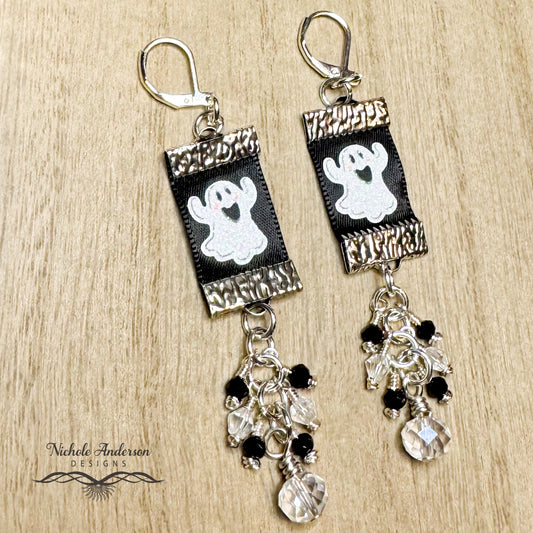 Halloween-themed ribbon ghost earrings with crystals suspended below shown on a wooden surface, featuring the brand 'Nichole Anderson Designs'.
