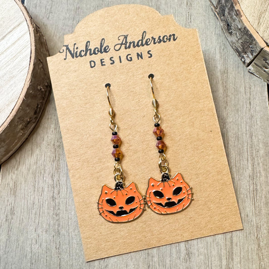 A pair of cat pumpkin earrings as jack o lanterns on an earring cards displayed as Nichole Anderson Designs