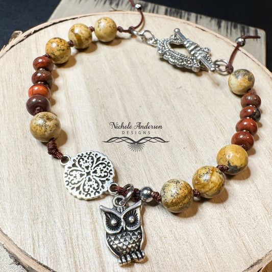 A knotted bracelet with red and picture jasper gemstones with a silver owl charm and medallion on a wooden surface, featuring 'Nichole Anderson Designs' branding.