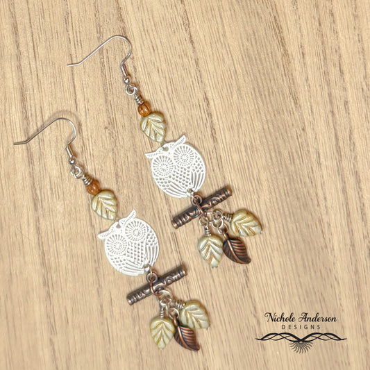 Pair of  silver owl-themed earrings with leaf charms on a wooden surface, featuring 'Nichole Anderson Designs' branding.