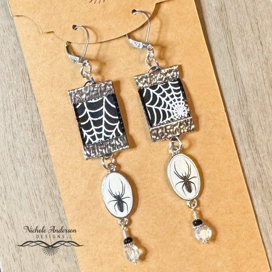 Spider web ribbon earrings with spider charm drop and black seed and crystal bead earrings on a brown card with 'Nichole Anderson Designs' branding.